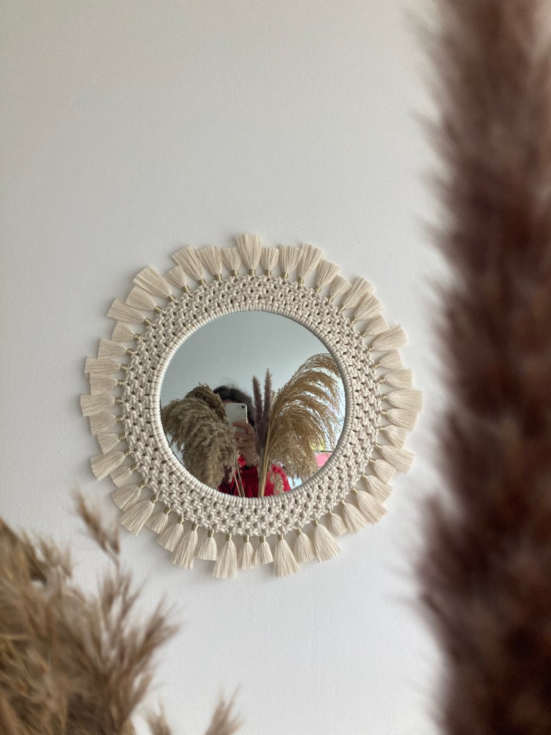 May include: A round mirror with a white macrame frame and tassels. The mirror is hanging on a white wall.