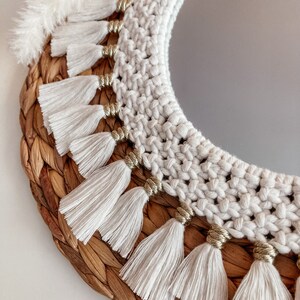 May include: A round mirror with a white crocheted frame and gold beads. The frame is decorated with white tassels. The mirror is set in a woven wicker base.