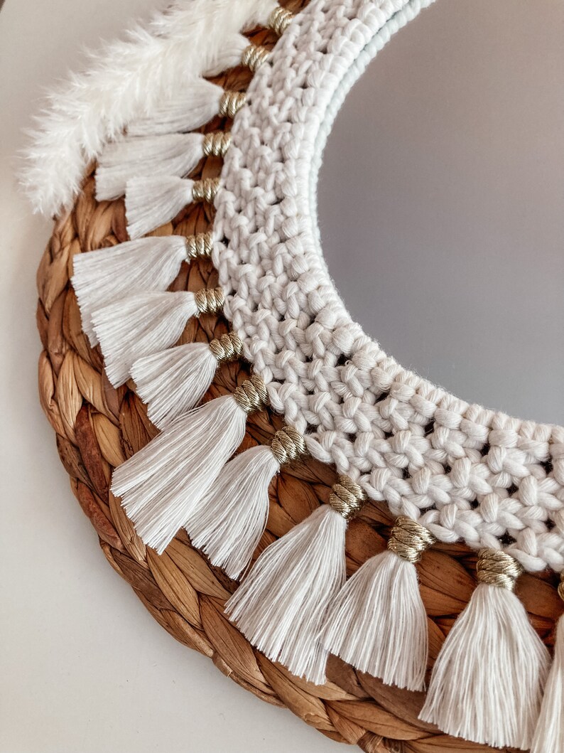 May include: A round mirror with a white crocheted frame and gold beads. The frame is decorated with white tassels. The mirror is set in a woven wicker base.