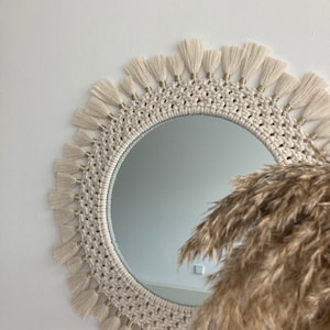 May include: A round macrame mirror with a white cotton cord and gold beads. The mirror has a fringe border and is hanging on a white wall.