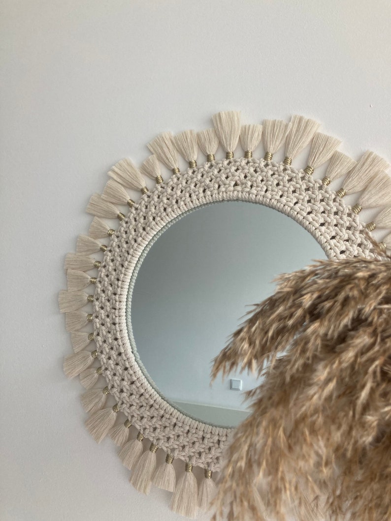 May include: A round macrame mirror with a white cotton cord and gold beads. The mirror has a fringe border and is hanging on a white wall.