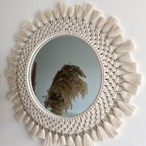 May include: A round macrame mirror with a white rope frame and gold beads. The mirror has a fringe border with white tassels.