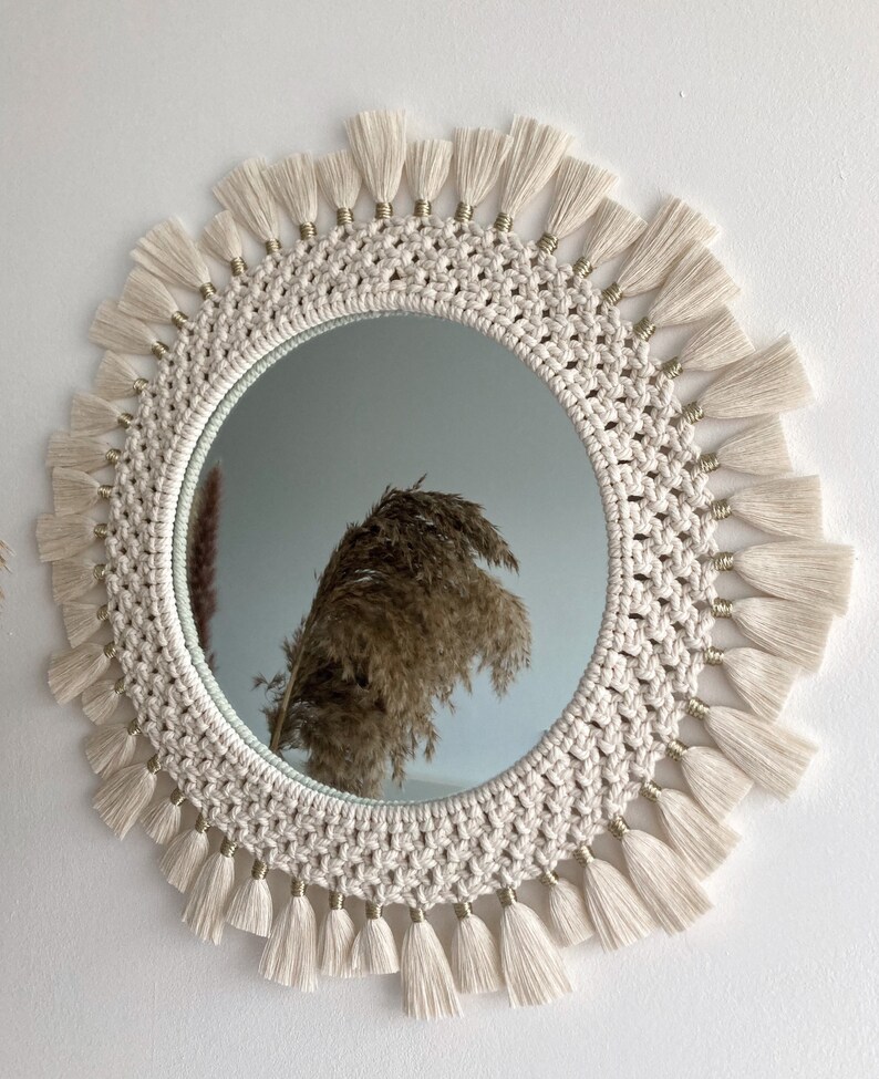May include: A round macrame mirror with a white rope frame and gold beads. The mirror has a fringe border with white tassels.