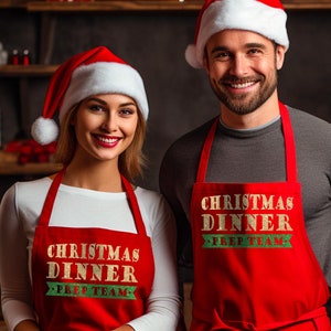 May include: Two red aprons with the text "Christmas Dinner Prep Team" in gold glitter lettering. The aprons are worn by two people wearing Santa hats.