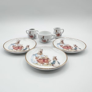 May include: A miniature porcelain tea set featuring three white saucers with gold rims, a small teacup, and two tiny pitchers. Each piece is decorated with illustrations of brown rabbits wearing blue jackets and red capes in a garden setting.