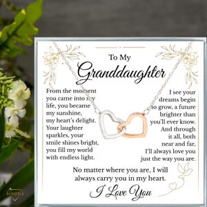 May include: A silver necklace with two interlocking heart pendants, one silver and one rose gold, on a white card. The card has text that reads "To My Granddaughter" and a loving message. The brand name "ROMESIA" is visible.