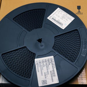 May include: A dark blue electronic component reel with a textured surface, labeled D330/16-100, sits inside a cardboard box. Two white labels with text and barcodes are affixed to the reel. The box has a fragile symbol.
