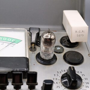 May include: Close-up of a vintage electronic device with a meter indicating "GOOD". A vacuum tube and a small box labeled "R.C.A: 5879" are visible. The device has black knobs and a silver metal surface.