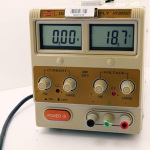 May include: A beige DC power supply unit with digital displays showing 0.00A and 18.7V. It has knobs for current and voltage adjustment, and a red power button. The unit has a black power cord.