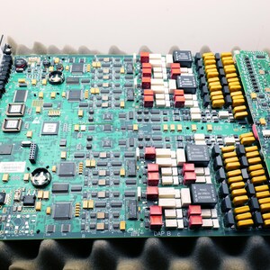 May include: A large green circuit board with numerous electronic components, including chips, resistors, and connectors. The board has a metal frame and is resting on a foam surface. Various colored components are visible.