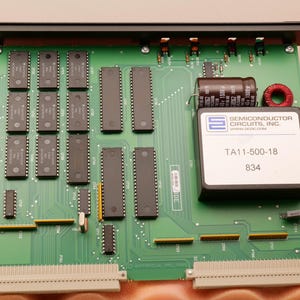 May include: A green circuit board with numerous black integrated circuits, capacitors, and connectors. A black box with the text "TA11-500-18" and "834" is visible. The board has a black metal frame.