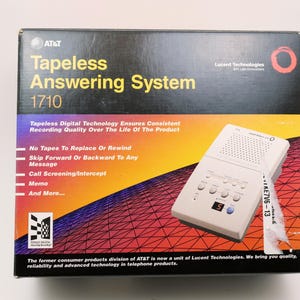 Puede incluir: Un sistema de contestador automático sin cinta AT&T 1710 en caja. La caja es negra con degradados amarillos y naranjas, y presenta el nombre y las características del producto. Se ve un contestador automático blanco. El texto incluye "No Tapes To Replace Or Rewind".