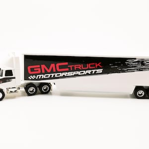 May include: A white GMC Truck Motorsports semi-truck toy. The truck has a white cab with black and red accents and a long trailer. The trailer features the words "GMC TRUCK MOTORSPORTS" in red and black.