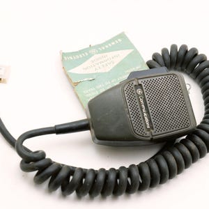 May include: A vintage General Electric Communications microphone with a coiled cord. The microphone is dark gray with a perforated speaker cover. A small connector and a safety information sheet are also present.