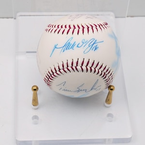May include: A white baseball with red stitching and multiple blue signatures. The baseball is displayed on a clear acrylic stand with two gold-colored supports. The baseball is a sports collectible.