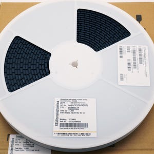 May include: A white plastic reel with dark gray components, labeled with text and barcodes, sits on a cardboard box. The reel is divided into sections, with the components filling the curved spaces. The label includes the text "STM Microelectronics".