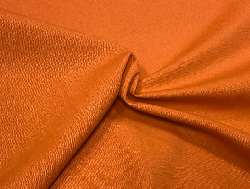 May include: A close-up of a rust-colored fabric with a smooth, woven texture. The fabric is folded and draped, showcasing its soft, pliable nature.