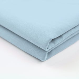May include: A stack of two folded, light blue fabric pieces. The fabric has a slightly textured appearance and a soft, muted color. The folds create a layered effect, highlighting the material's drape.