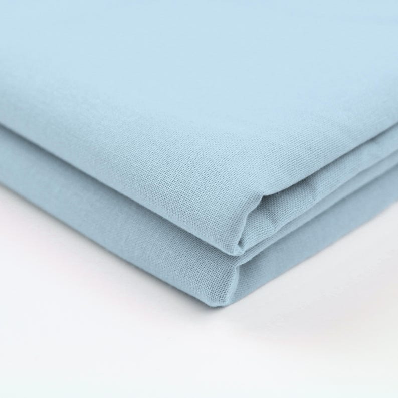 May include: A stack of two folded, light blue fabric pieces. The fabric has a slightly textured appearance and a soft, muted color. The folds create a layered effect, highlighting the material's drape.
