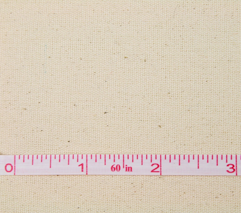 May include: A close-up of a plain, off-white canvas fabric with a pink measuring tape laid across it. The tape reads "60 in" and has markings for 0, 1, 2, and 3 inches.