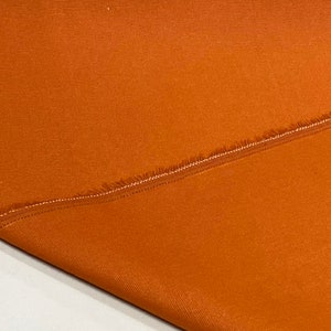 May include: A close-up of a folded piece of burnt orange fabric with a raw edge. The fabric has a subtle texture and a slight sheen.