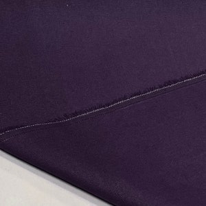 May include: A close-up of a dark purple fabric with a raw edge and a single stitch line. The fabric has a subtle texture.
