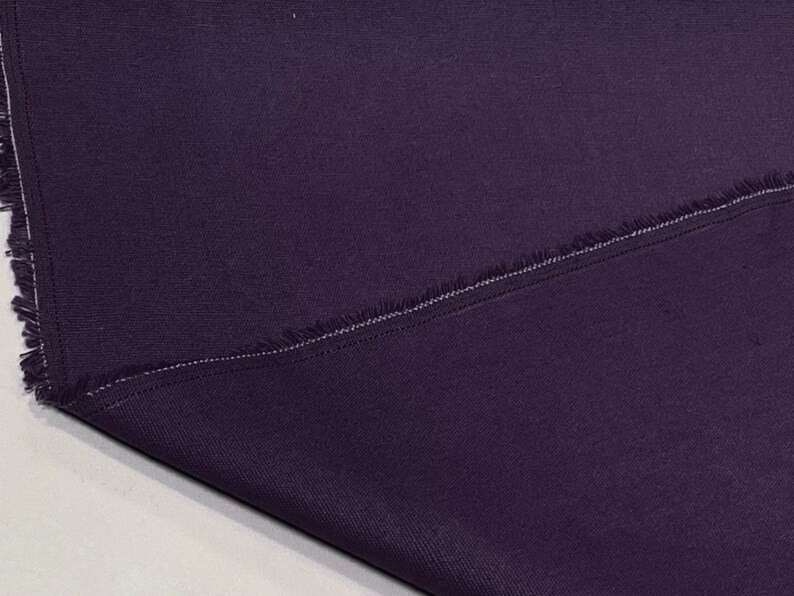May include: A close-up of a dark purple fabric with a raw edge and a single stitch line. The fabric has a subtle texture.