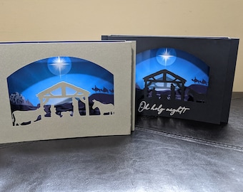 May include: Two Christmas card pop-up scenes with a blue night sky, a star, and a nativity scene. The card on the left is gray and the card on the right is black. The black card has the text "Oh holy night" written on it.