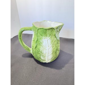 May include: A ceramic pitcher shaped like a head of cabbage, featuring a textured green and white leaf pattern with a prominent central vein and a curved handle. The interior is smooth and white, while the exterior mimics the appearance of layered cabbage leaves with realistic veining and a glossy finish.