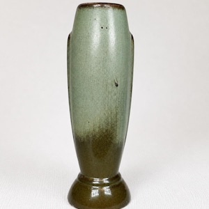 May include: A tall ceramic vase featuring a smooth, tapered cylindrical body with a flared circular base. The surface displays a two-tone glaze, transitioning from a muted sage green at the top to a deep, earthy brown at the bottom and rim.