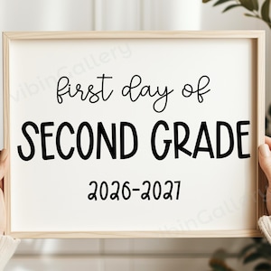 Può includere: Un cartello rettangolare in una cornice di legno chiaro con testo nero su sfondo bianco. Il testo recita first day of SECOND GRADE 2026-2027. Il cartello è tenuto da due mani che indossano maglioni beige a maniche lunghe su sfondo sfocato.