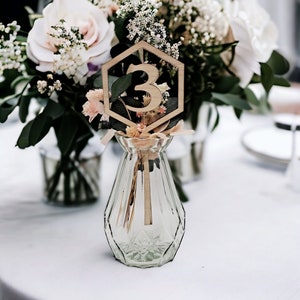 May include: A wooden table number with a geometric hexagon shape and the number "3" in the center. The table number is placed in a clear glass vase with dried flowers.