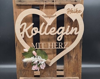 May include: A wooden heart-shaped sign with the text "Heike Kollegin mit Herz" hanging on a wooden crate. The sign is decorated with a small bouquet of dried flowers.