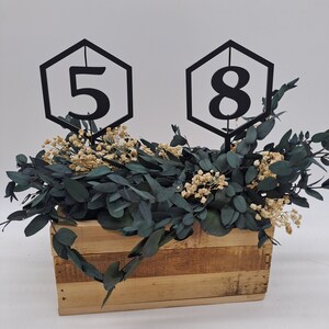 May include: Two black hexagonal table numbers, one with the number "5" and the other with the number "8", are placed in a wooden box filled with green eucalyptus and white flowers.