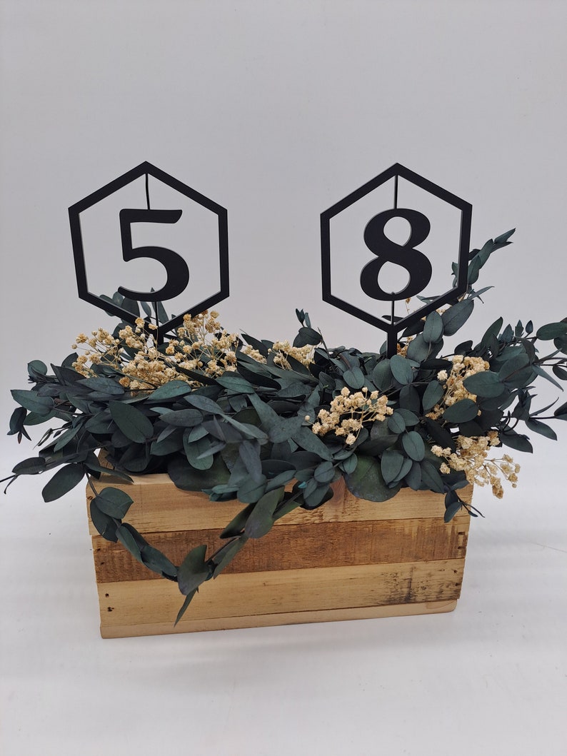 May include: Two black hexagonal table numbers, one with the number "5" and the other with the number "8", are placed in a wooden box filled with green eucalyptus and white flowers.