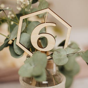 May include: A wooden table number six in a hexagonal frame, placed in a clear glass vase wrapped in twine, with green eucalyptus leaves.