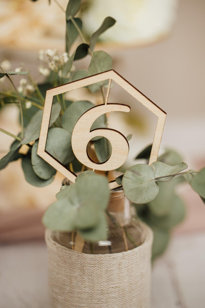 May include: A wooden table number six in a hexagonal frame, placed in a clear glass vase wrapped in twine, with green eucalyptus leaves.