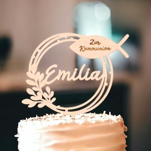 Pode incluir: Topper de bolo de madeira com um c&iacute;rculo e um peixe com o texto "Zur Kommunion" e o nome "Emilia" em it&aacute;lico.