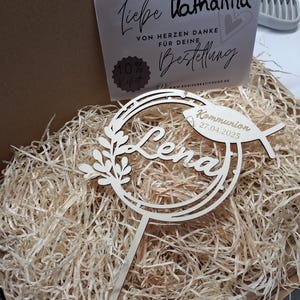 Pode incluir: Topper de bolo de madeira com um design circular e o nome "Lena" em it&aacute;lico. O topper tamb&eacute;m inclui um pequeno peixe com o texto "Kommunion 27.04.2025".