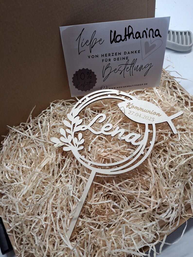 Pode incluir: Topper de bolo de madeira com um design circular e o nome "Lena" em it&aacute;lico. O topper tamb&eacute;m inclui um pequeno peixe com o texto "Kommunion 27.04.2025".