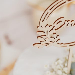 Pode incluir: Um letreiro de madeira com a palavra "Emma" esculpida nele. O letreiro &eacute; decorado com um desenho de folhas e est&aacute; preso a um pau de madeira.