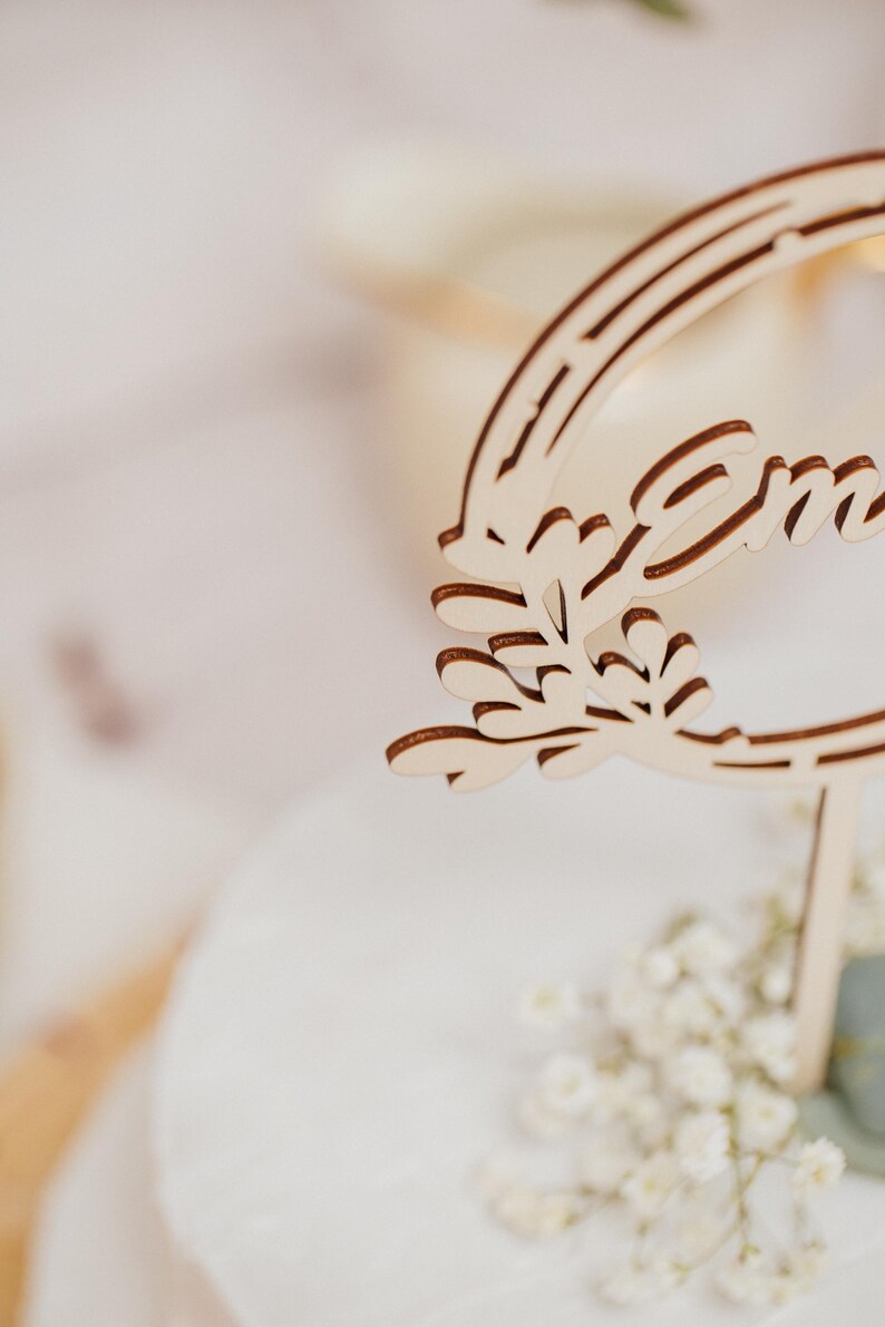 Pode incluir: Um letreiro de madeira com a palavra "Emma" esculpida nele. O letreiro &eacute; decorado com um desenho de folhas e est&aacute; preso a um pau de madeira.