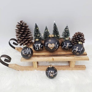 Pode incluir: Seis bolas de Natal pretas com letras douradas em um tren&oacute; de madeira. As bolas s&atilde;o decoradas com nomes, incluindo "Tyler Family", "Kristof", "Joshua", "Janet" e "Taco" com uma pegada de pata. O tren&oacute; &eacute; cercado por &aacute;rvores de pinho em miniatura e pinhas.