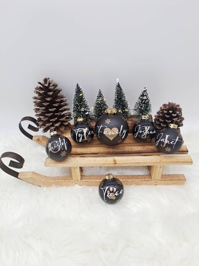 Pode incluir: Seis bolas de Natal pretas com letras douradas em um tren&oacute; de madeira. As bolas s&atilde;o decoradas com nomes, incluindo "Tyler Family", "Kristof", "Joshua", "Janet" e "Taco" com uma pegada de pata. O tren&oacute; &eacute; cercado por &aacute;rvores de pinho em miniatura e pinhas.