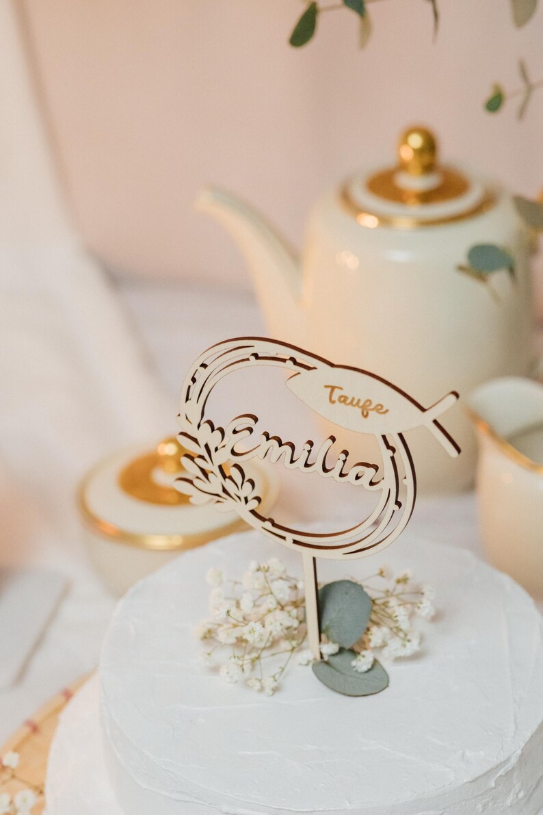 Pode incluir: Um bolo branco com um topo de bolo de madeira que diz "Taufe Emilia". O topo &eacute; decorado com um pequeno p&aacute;ssaro e folhas.