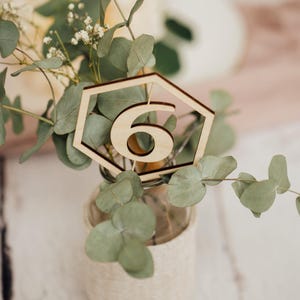 May include: A wooden table number six in a hexagonal shape, placed in a clear glass jar with a burlap ribbon around it. The jar is filled with green eucalyptus leaves.