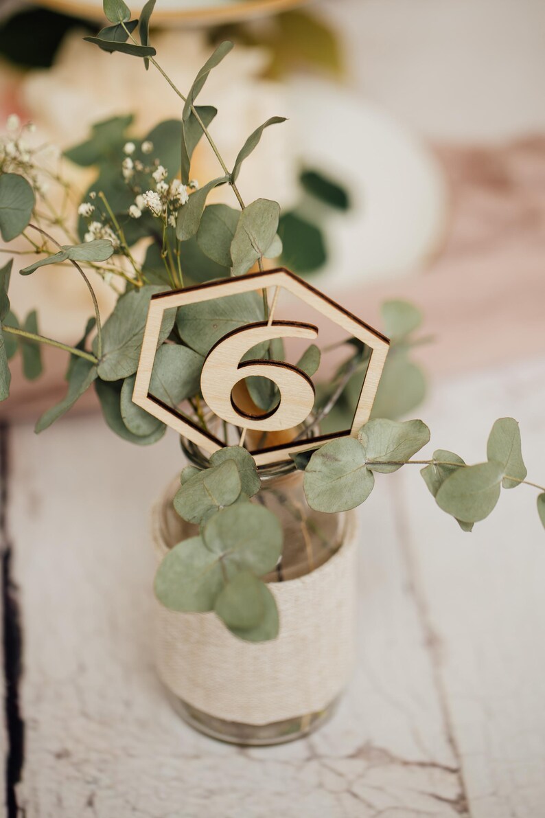 May include: A wooden table number six in a hexagonal shape, placed in a clear glass jar with a burlap ribbon around it. The jar is filled with green eucalyptus leaves.