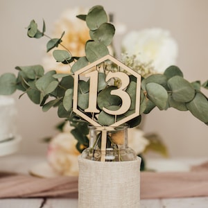May include: Wooden table number with the number 13 in a hexagonal shape, placed in a clear glass vase with a burlap wrap, surrounded by greenery.