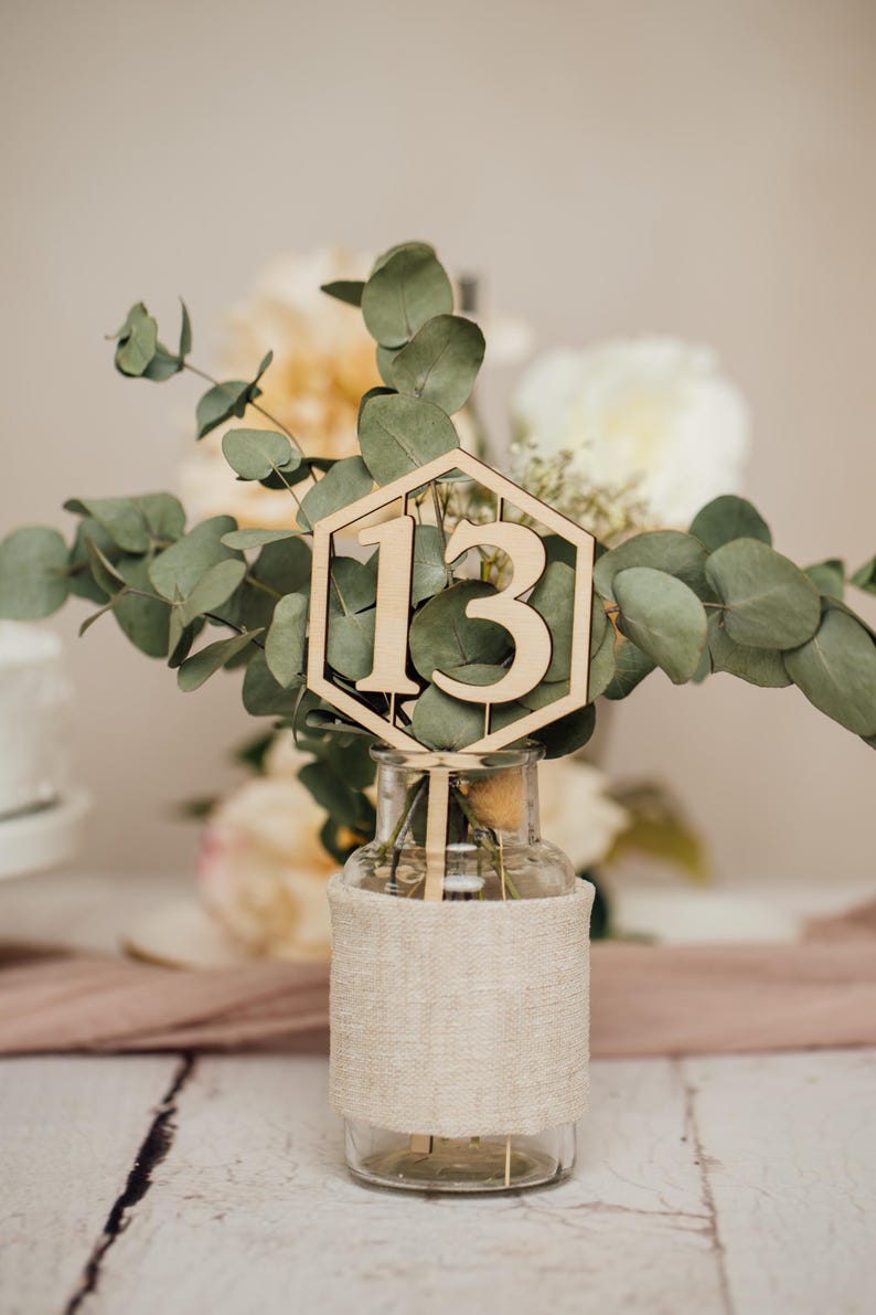 May include: Wooden table number with the number 13 in a hexagonal shape, placed in a clear glass vase with a burlap wrap, surrounded by greenery.