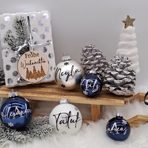 Pode incluir: Um conjunto de seis enfeites de Natal personalizados com detalhes prateados e azuis. Os enfeites s&atilde;o decorados com nomes e flocos de neve. Os enfeites s&atilde;o exibidos em um tren&oacute; de madeira com uma base de pele sint&eacute;tica branca. Os enfeites s&atilde;o cercados por pinhas e uma pequena &aacute;rvore de Natal artificial branca.